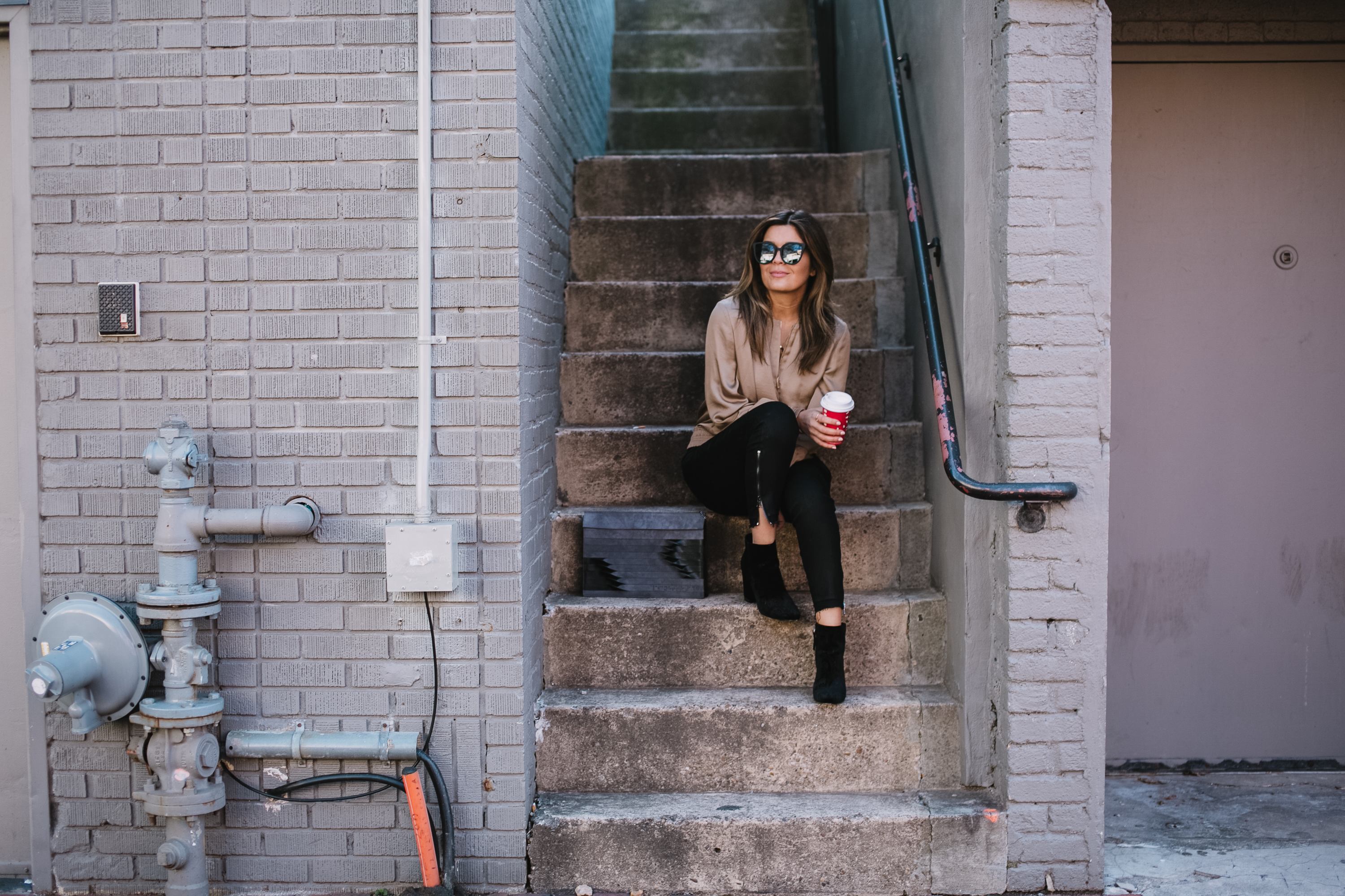 Blogger Sara Azani of Style MBA is photographed wearing Lafayette 148 New York Bryant satin bomber jacket, Quay Australia black oversize sunglasses, J Brand Coated Skinny Jeans, Target paisley booties, and limited edition ITA for Lafayette 148 New York Shantou Pouchette purse. Petite brunette woman wearing simple neutral outfit holding a small coffee while sitting in an outdoor stairwell.