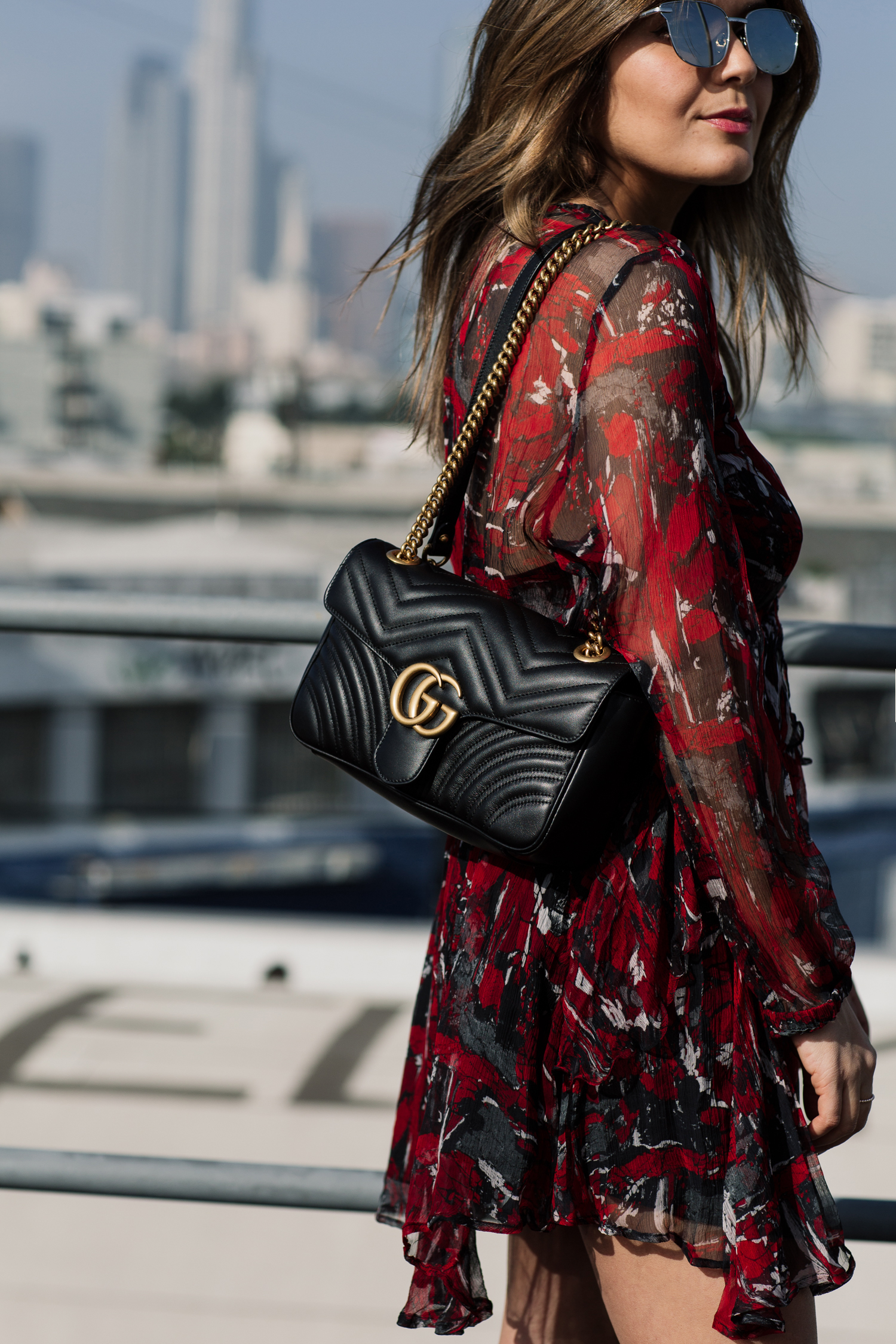 Brunette woman, blogger Sara Azani of Style MBA poses outside in front of a cityscape during the daytime wearing an IRO little red dress, Gucci GG Marmont quilted leather shoulder bag with gold hardware, Chloé Susanna Stud Buckle Booties.